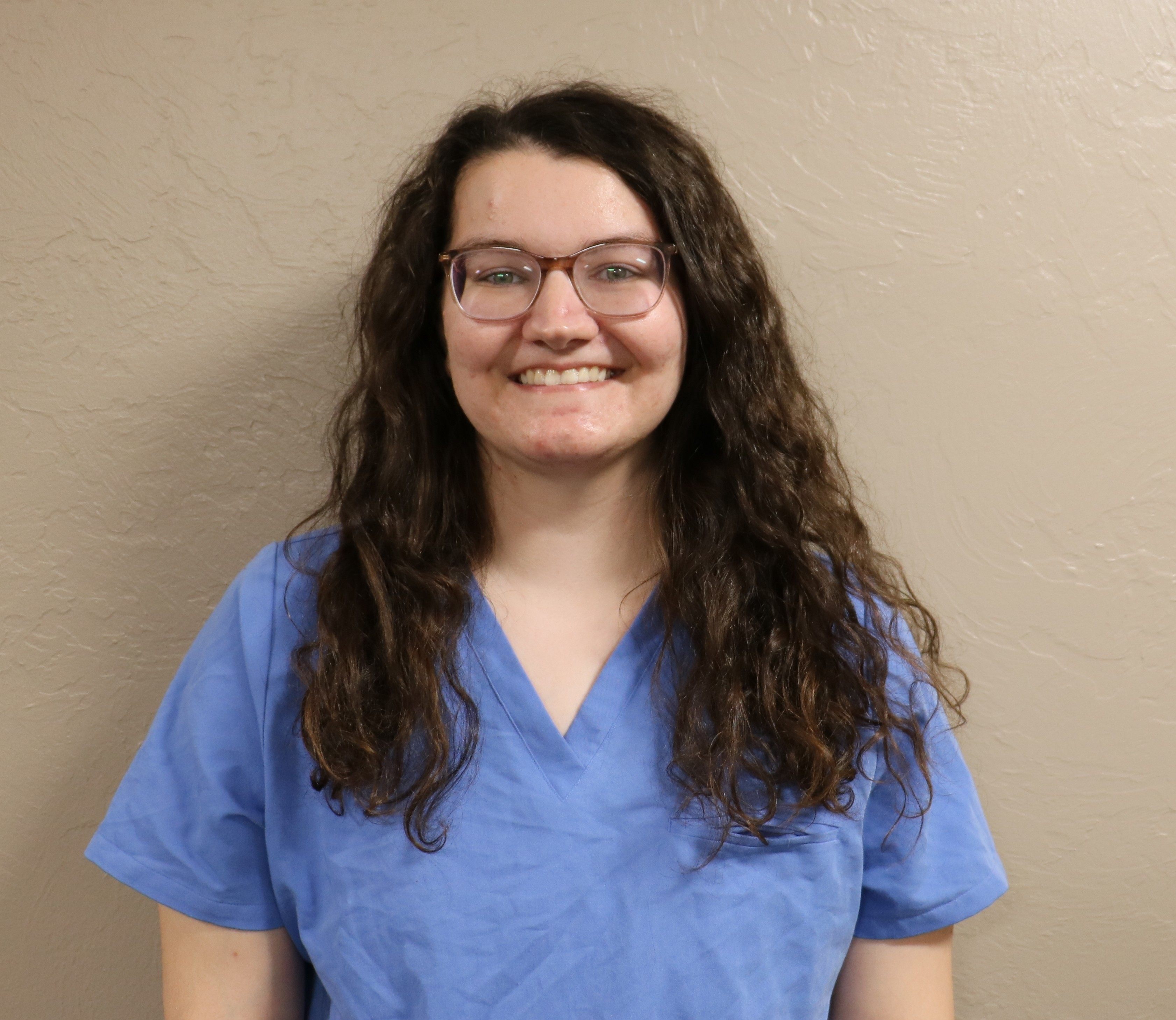 Bradie Frizzell Photo of a woman in blue hospital scrubs with long dark hair and wearin glasses smilig