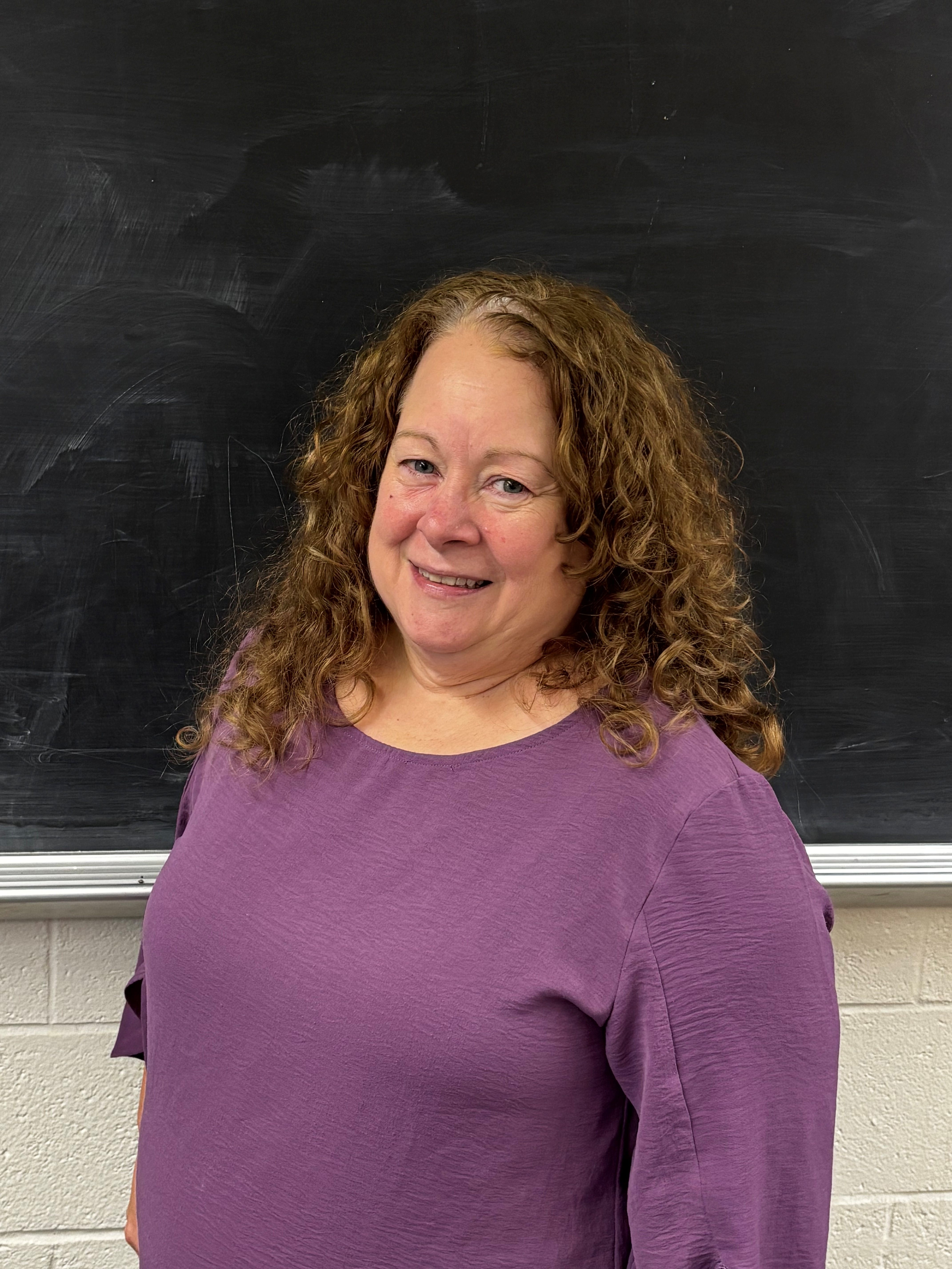 Portrait of a woman with long curly hair smiling in front of a blakcboard, wearing a purple shirt